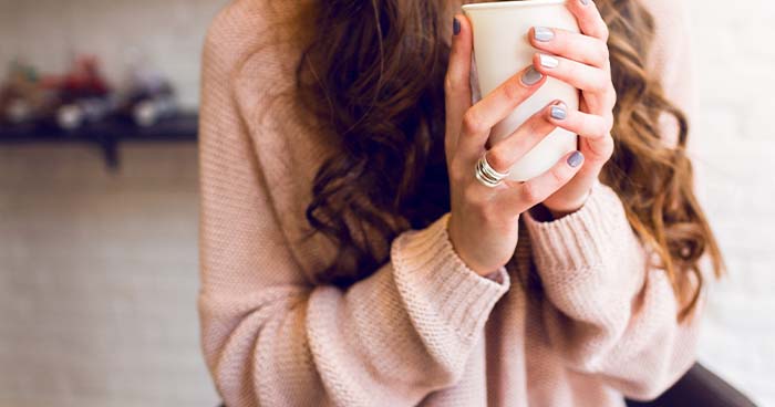 Image: a woman holds a ceramic mug close to her chest. Wearing sweaters and actually dressing for the colder weather can help lower your electric bills.