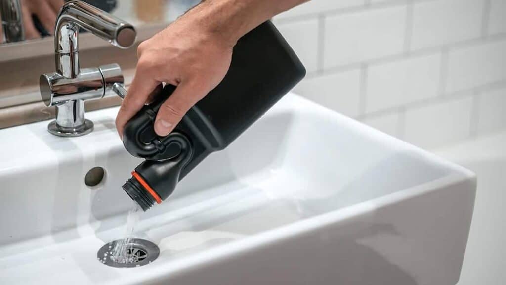 An Adeedo technician carefully pours cleaning solution from a black bottle into a white bathroom sink drain.
