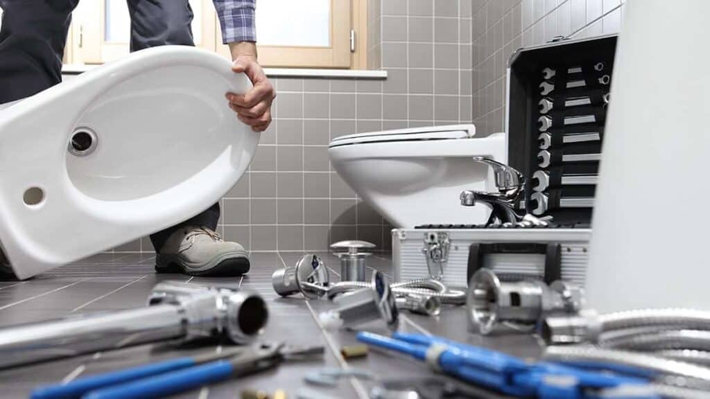 An Adeedo technician installs a bathroom sink, with plumbing tools and parts on a tiled floor next to a toilet and tool kit.