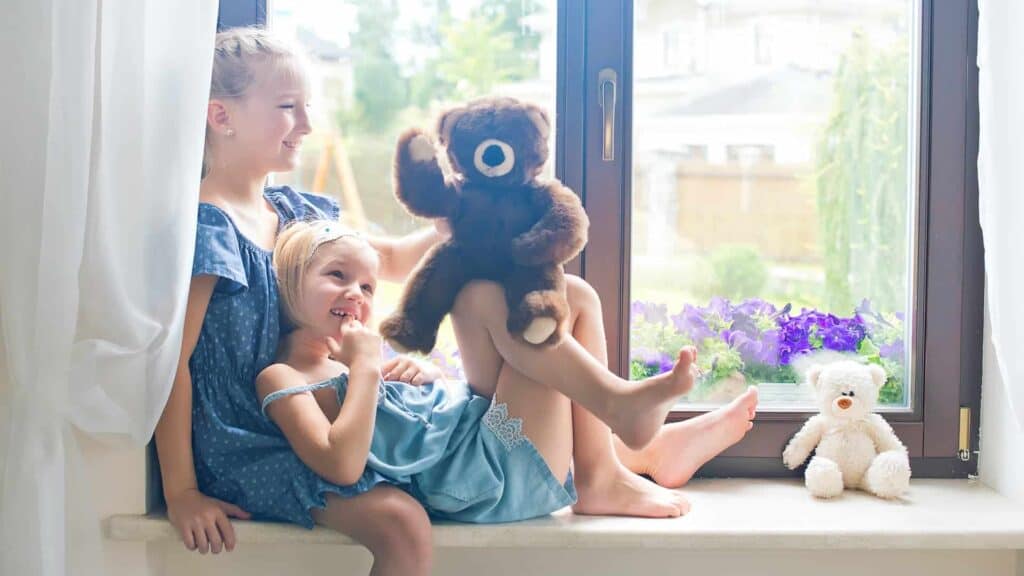 Two young Adeedo customers sit on a windowsill—one holding a brown teddy bear, with a white teddy bear and flowers nearby.