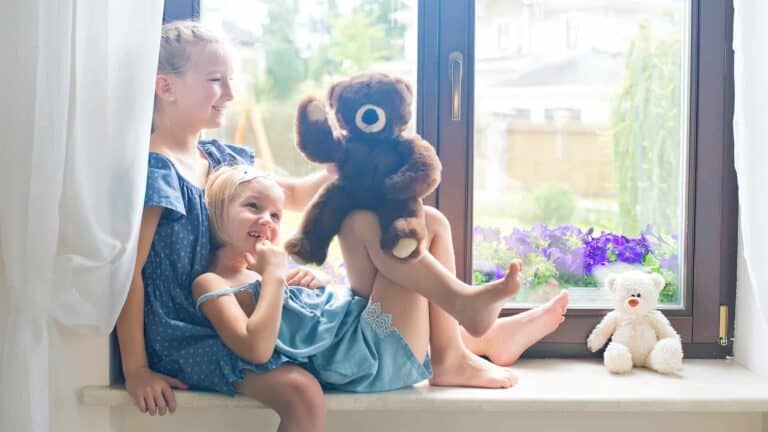 Two young Adeedo customers sit on a windowsill—one holding a brown teddy bear, with a white teddy bear and flowers nearby.