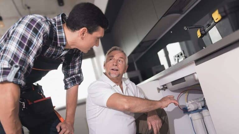 An Adeedo technician and customer review plumbing under a kitchen sink, discussing solutions in a friendly, professional manner.