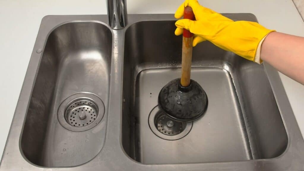 An Adeedo team member in yellow gloves uses a plunger to clear a stainless steel kitchen sink, ensuring top-quality service.
