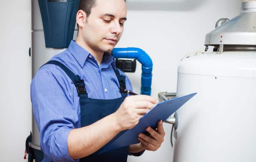 An Adeedo technician in a blue shirt and overalls reviews a clipboard while inspecting a water heater and plumbing system.