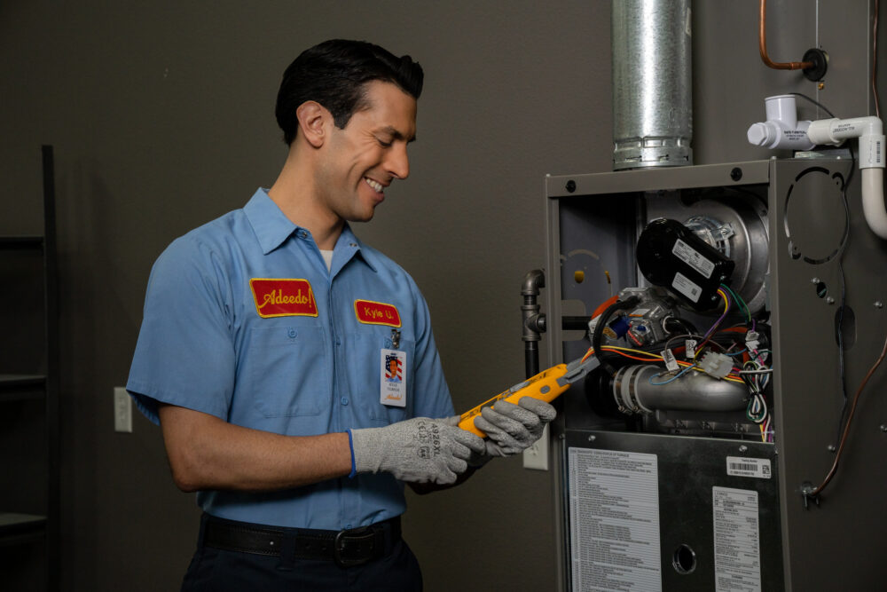 An Adeedo technician, dressed in a blue uniform, uses a yellow multimeter to carefully inspect the wiring inside an open furnace. Smiling, he ensures the heating system is safe and ready for winter, providing reliable service to Southern California customers.